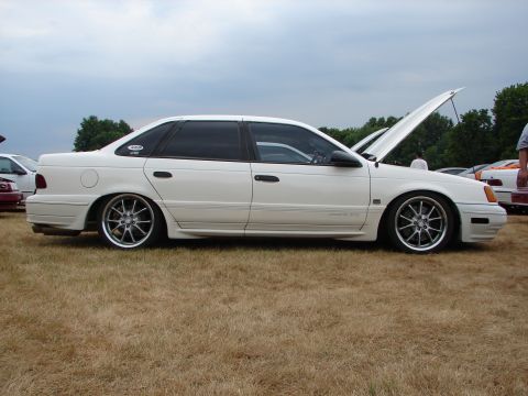 1993 Ford Taurus - A white 1993 Ford Taurus sedan is displayed with its hood open and lowered suspension at a car show.