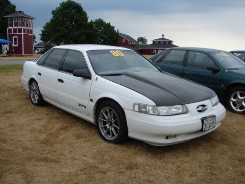 1993 Ford Taurus - A white 1993 Ford Taurus with a black hood is parked on dry grass next to another car.