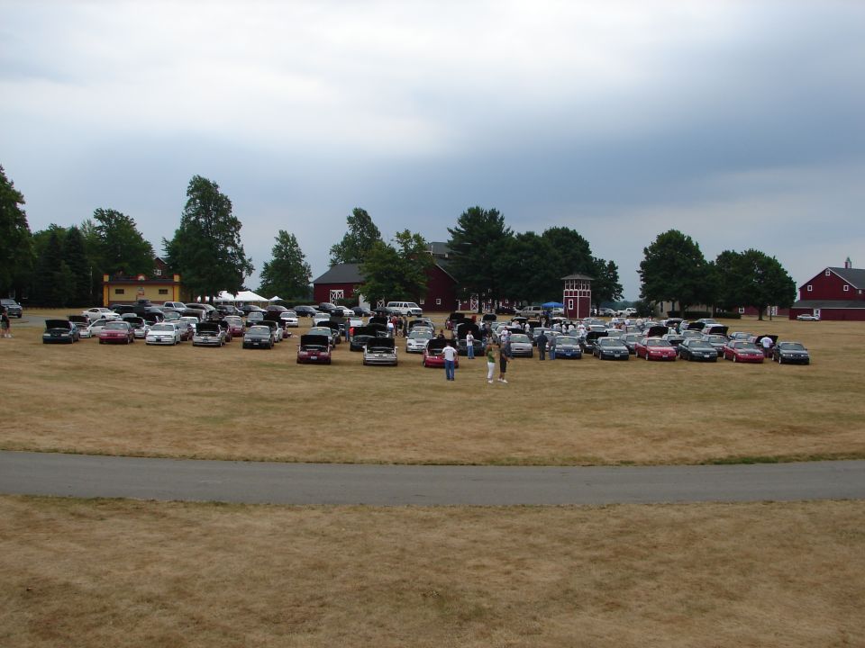 Car show scene with numerous vehicles parked on dry grass, some with hoods open