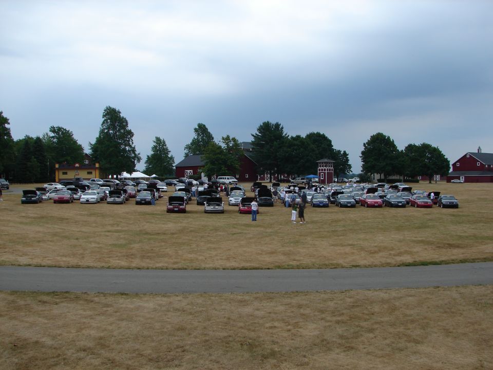 Many cars parked on a dry grass field with buildings and trees in the background under a cloudy sky
