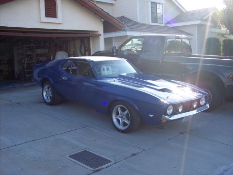 1971 Ford Mustang - A blue 1971 Ford Mustang with a prominent hood scoop and aftermarket wheels is parked in front of a house and a dark pickup truck.