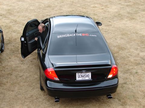 1993 Ford Taurus - Black sedan with a custom sticker and license plate parked on dry grass.