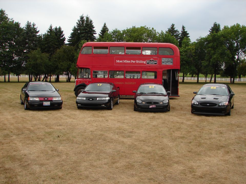 Four black Ford Taurus cars parked in front of a red double-decker bus