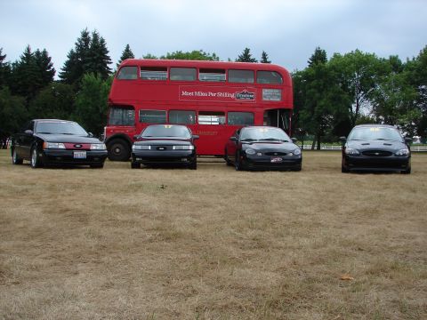 1993 Ford Taurus - Four black 1993 Ford Taurus sedans are lined up in front of a red double-decker bus on a dry, grassy field.
