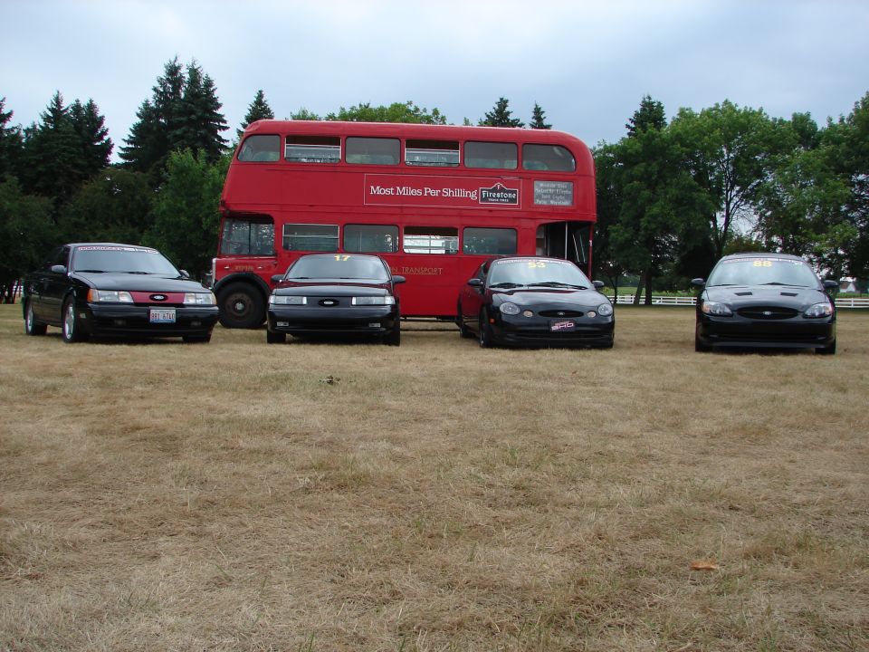 Four black 1993 Ford Taurus sedans parked in front of a red double-decker bus