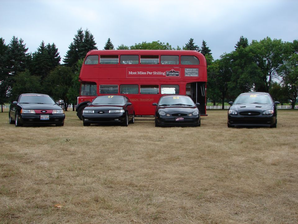 Four black Ford Taurus sedans parked in front of a red double-decker bus on a dry grass field