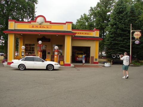 1993 Ford Taurus - A white 1993 Ford Taurus is parked in front of a restored Shell gas station, with a vintage truck visible inside the garage.