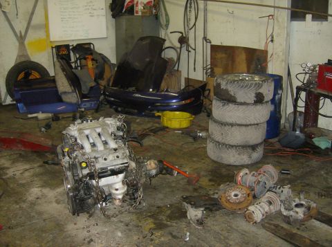 1996 Ford Probe - Various car parts, including an engine and tires, are piled up in a cluttered garage space.