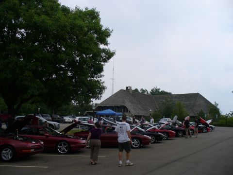 1991 Ford Probe - A gathering of cars, mostly red coupes, with their hoods open, parked in a lot near a building.