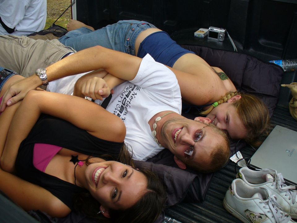Three people are lying down in the back of a truck bed. One person is wearing a white t-shirt with text on it