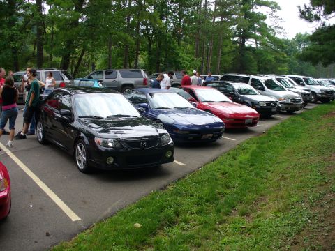1991 Ford Probe - A row of cars are parked in a lot, with people standing near the front vehicles.