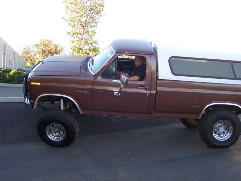 1991 Ford Bronco - A lifted brown pickup truck with a white camper shell is parked outside on a paved surface.