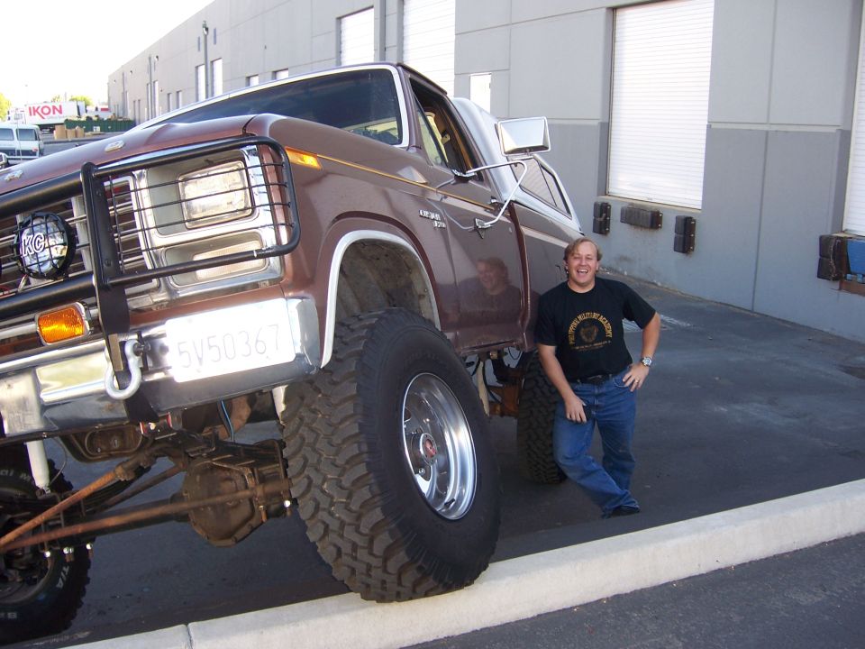 Lifted brown Ford pickup truck with large tires, bull bar, and KC lights. Man stands next to it. License plate reads 5V50367