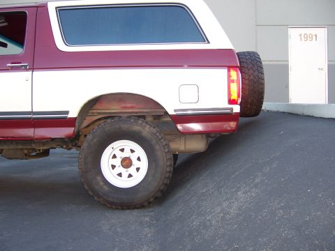 1991 Ford Bronco - The rear of a two-tone Ford Bronco is visible, with a spare tire mounted on the back. A door with the number 1991 is in the background.