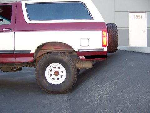 1991 Ford Bronco - The rear of a two-tone SUV is shown, with a spare tire mounted on the back. A building with the number 1991 above its door is visible in th…