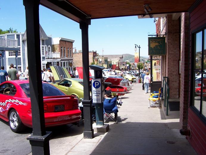 Street lined with parked cars, some with hoods up, in front of shops. A person sits in a chair near a parking meter