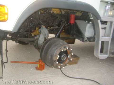 1990 Ford Bronco - truck's front suspension and exposed brake hub with the wheel removed, leaf spring and shock visible, with an orange bottle jack underneath.