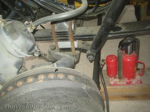 1990 Ford Bronco - truck's front brake hub and leaf-spring suspension with a red hydraulic bottle jack resting on a wooden block underneath.
