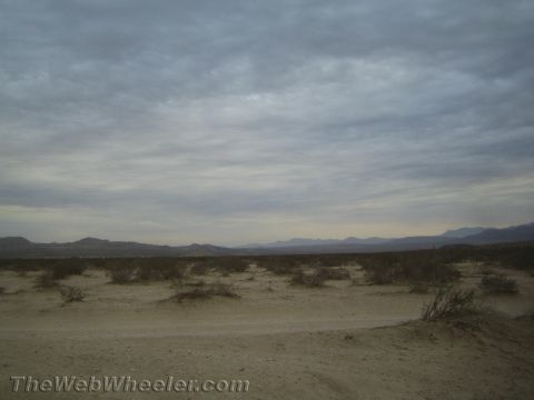 1990 Ford Bronco - flat desert plain with sparse brush and a low mountain range on the horizon beneath a cloudy sky.