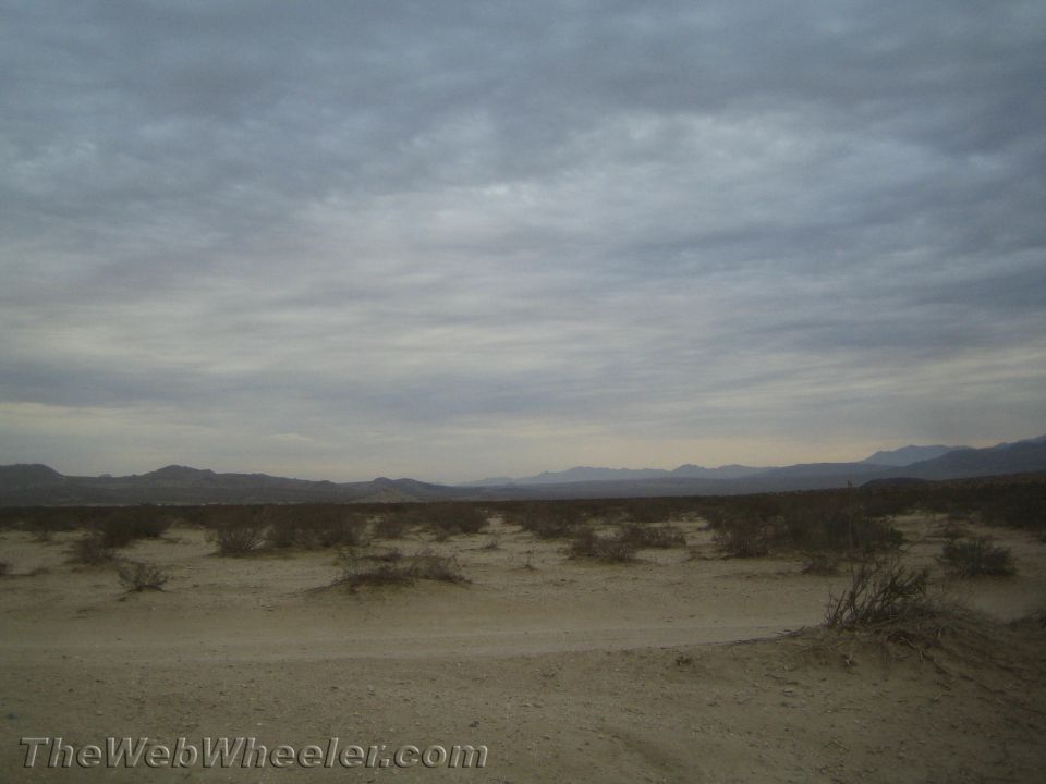 Flat desert plain with sparse brush and mountains under a cloudy sky