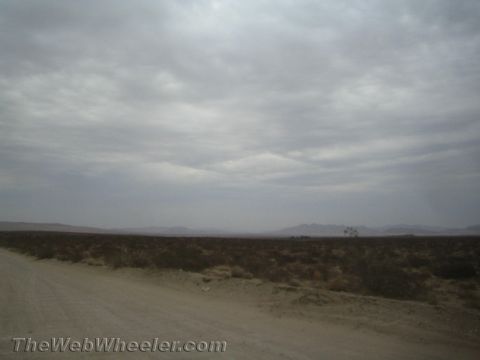 1990 Ford Bronco - dirt road runs alongside desert scrubland, with hazy mountains in the distance under a heavily overcast sky.