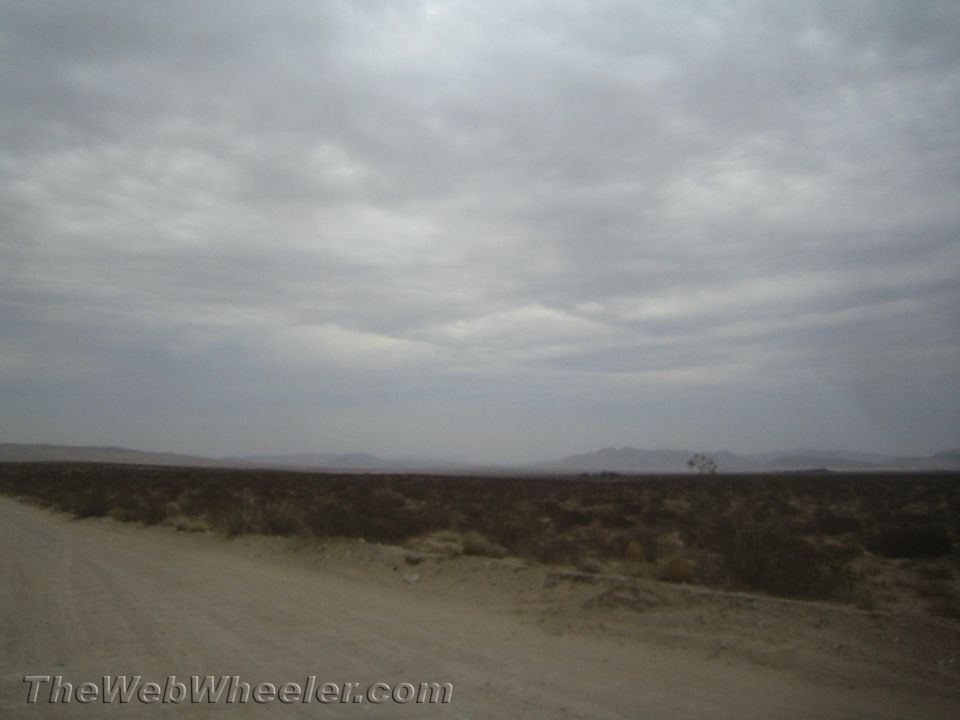 Desert dirt road beside scrubland with hazy mountains under overcast sky