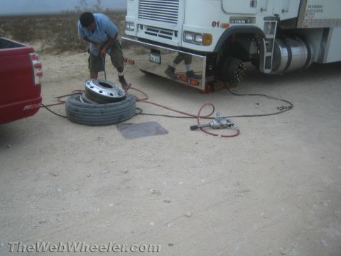 1990 Ford Bronco - man works on a large truck wheel lying on the desert ground beside a white commercial truck, with air hoses and tools spread out.
