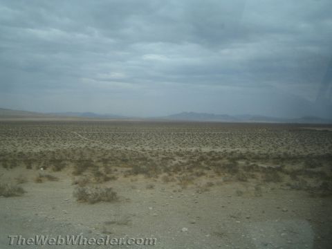 1990 Ford Bronco - wide, flat desert plain dotted with sparse scrub brush stretches toward distant hazy mountains beneath a cloudy gray sky.