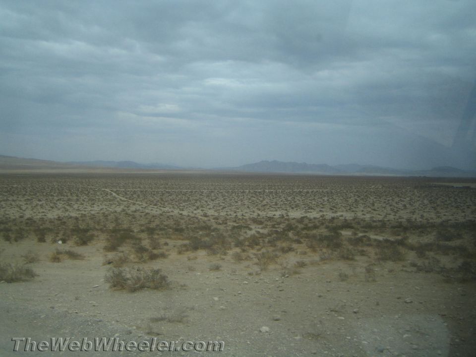 Flat desert landscape with sparse brush under an overcast gray sky and distant mountains