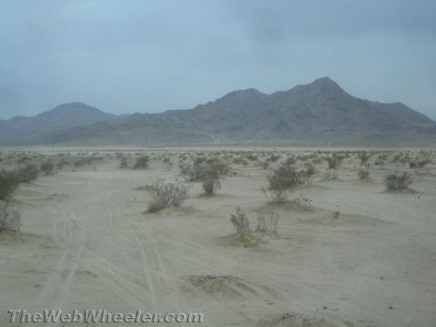 1990 Ford Bronco - sandy desert flat scattered with low shrubs and faint tire tracks sits in front of a rugged mountain ridge under hazy sky.