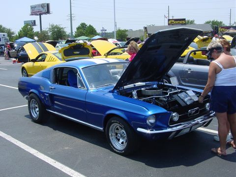 1986 Ford Mustang - A blue 1967 Ford Mustang coupe with white racing stripes is displayed at an outdoor car show.