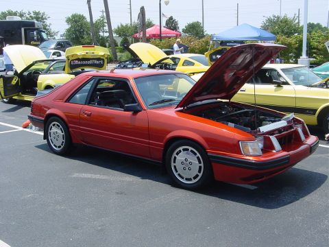 1986 Ford Mustang - This orange 1986 Ford Mustang coupe is displayed with its hood open at a car show, alongside other vehicles.