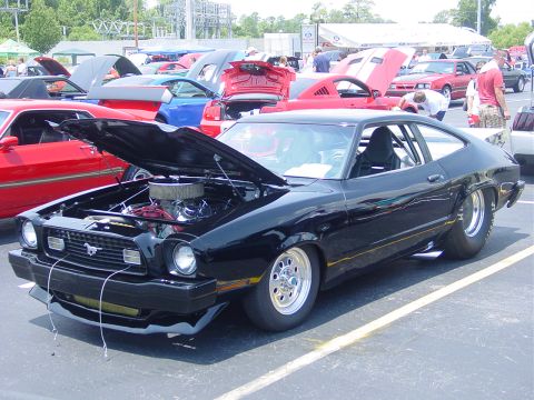 1986 Ford Mustang - A black coupe with its hood open is displayed at an outdoor car show with other vehicles around.