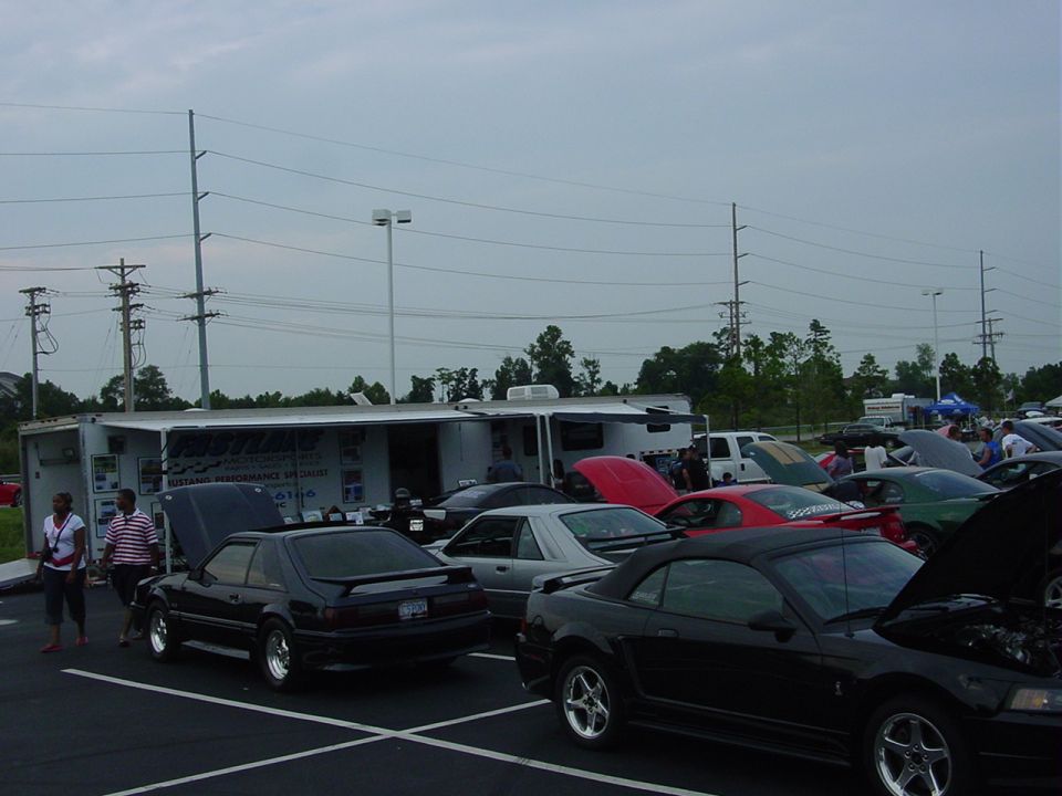 Several Ford Mustangs parked outside a trailer with hoods up. Some people are standing around