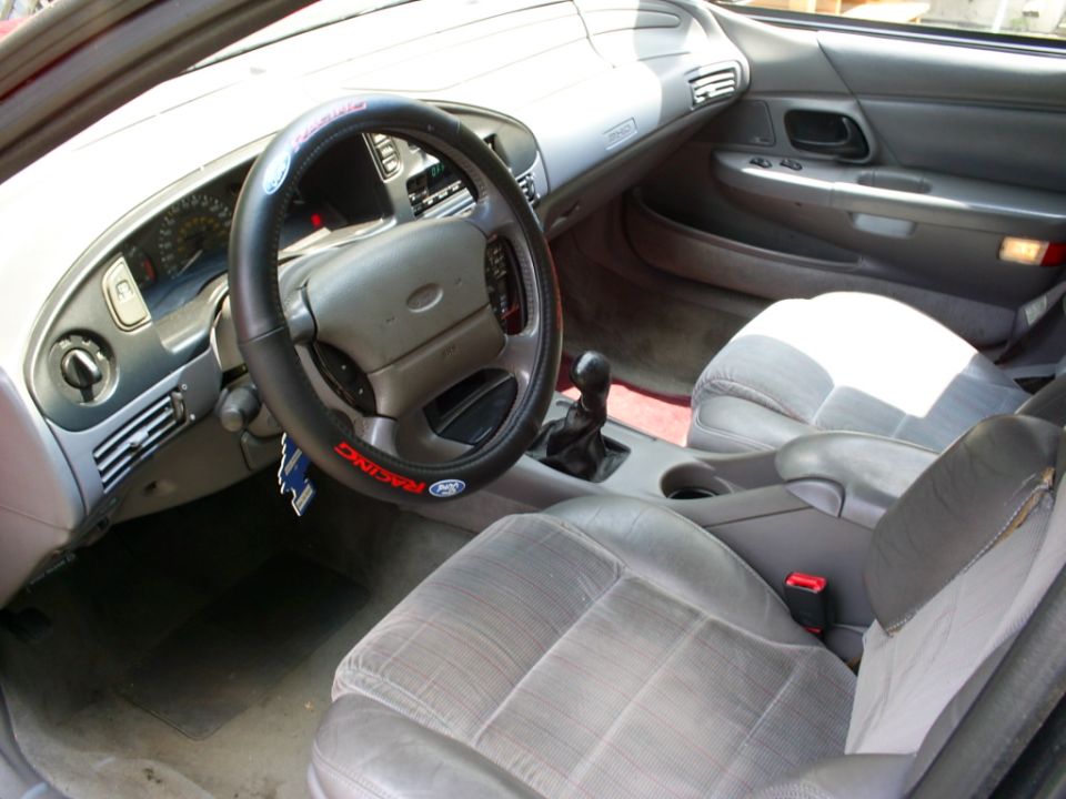 Interior shot of a 1995 Ford Taurus, showing the dashboard, steering wheel with a racing cover, and front seats