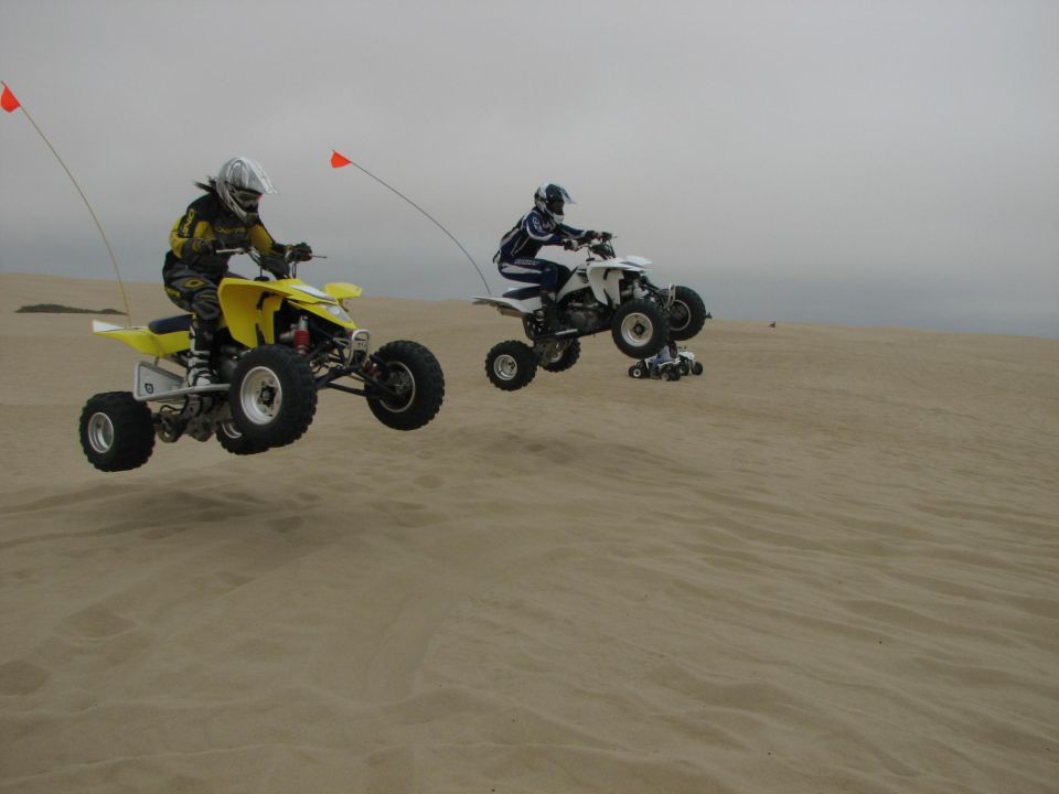 Two riders jumping sport ATVs mid-air over sand dunes, yellow quad leading a white quad