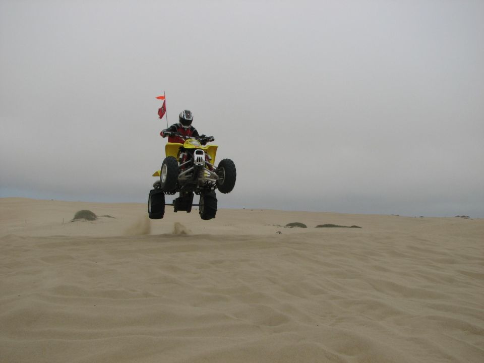 Rider jumping a yellow ATV with orange whip flag over sand dunes under overcast sky