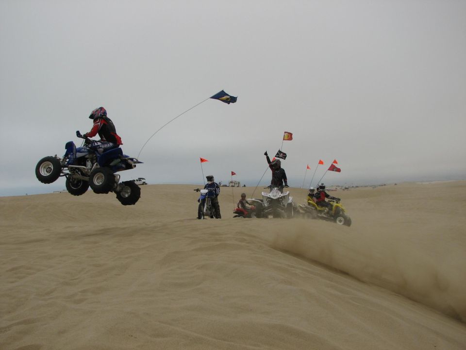 Rider jumps a blue Yamaha ATV over a sand dune as other quads and a dirt bike with whip flags watch below