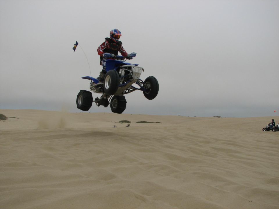 Blue sport ATV with paddle tires airborne over sand dunes, rider in red-white-blue helmet