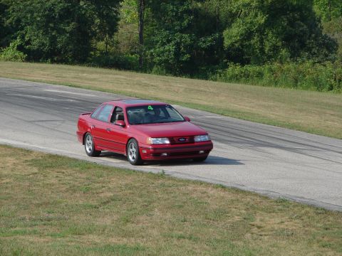 1993 Ford Taurus - A red sedan, likely a 1992 model, navigates a turn on a road course, indicated by tire marks on the asphalt.