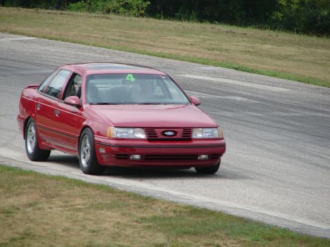 1993 Ford Taurus - A red sedan navigates a turn on a paved race track, with grass and trees visible in the background.