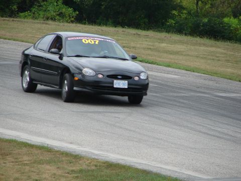 1993 Ford Taurus - A black sedan navigates a turn on a race track, displaying racing numbers on its windshield.
