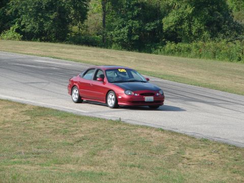 1993 Ford Taurus - Red sedan with a black hood and racing number 08 on the windshield drives on a paved road course.