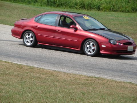 1993 Ford Taurus - A red sedan with a black hood navigates a road course, displaying a yellow '00' sticker on its windshield.