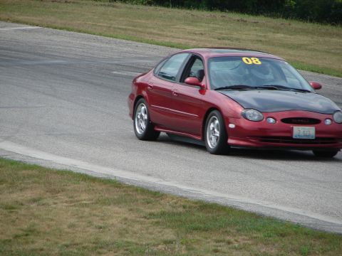 1993 Ford Taurus - A red sedan with a black hood and yellow racing number 08 on the windshield navigates a turn on a road course.