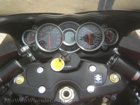 1990 Ford Bronco - Close-up of a Suzuki motorcycle's instrument cluster showing twin analog gauges, a yellow-tagged key in the ignition, and the Suzuki logo.