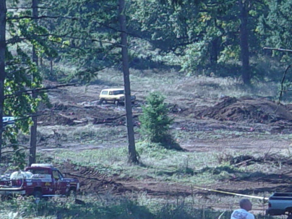 Yellow SUV in a distant clearing seen through trees, red pickup parked at lower left