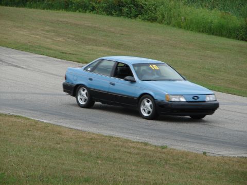 1993 Ford Taurus - Blue 1993 Ford Taurus driving on a road course with grass and trees in the background.