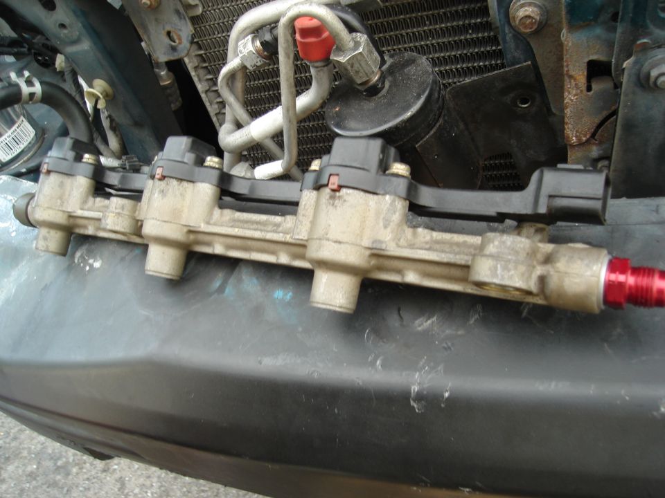 Close-up of a dirty fuel rail with red fittings, connected to engine components and a radiator