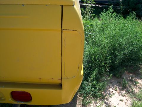1979 Ford Bronco - Close-up of the rear quarter panel of a yellow vehicle, showing a red taillight and overgrown green foliage.